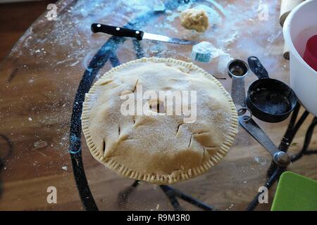 Unbaked apple pie on messy glass table surrounded by measuring spoons, cup, and knife. Stock Photo