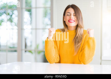 Young beautiful woman wearing winter sweater at home excited for success with arms raised celebrating victory smiling. Winner concept. Stock Photo