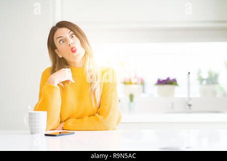 Young beautiful woman drinking a cup of coffee at home making fish face with lips, crazy and comical gesture. Funny expression. Stock Photo