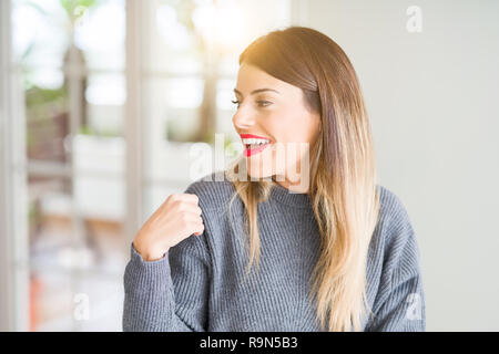 Young beautiful woman wearing winter sweater at home looking away to side with smile on face, natural expression. Laughing confident.