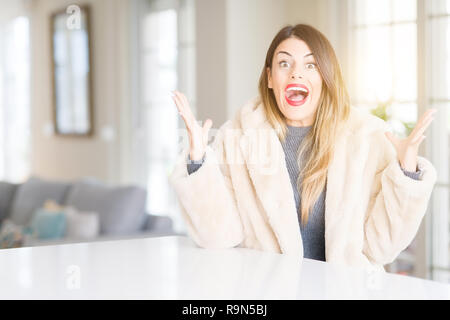 Young beautiful woman wearing winter fur coat at home celebrating crazy and amazed for success with arms raised and open eyes screaming excited. Winne Stock Photo