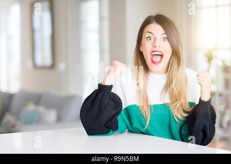 Young beautiful woman wearing winter sweater at home celebrating surprised and amazed for success with arms raised and open eyes. Winner concept. Stock Photo