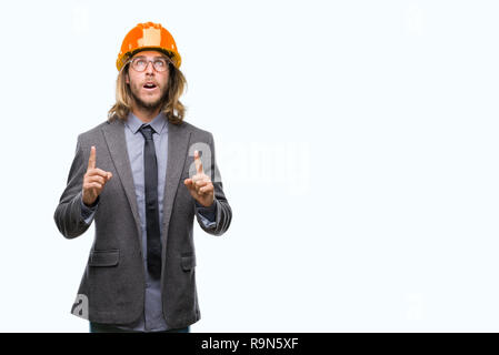 Young handsome architec man with long hair wearing safety helmet over isolated background amazed and surprised looking up and pointing with fingers an Stock Photo