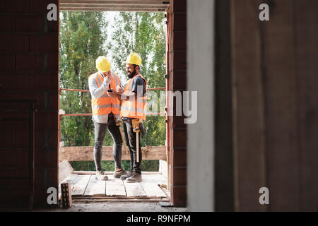 Persons working in construction site. Latino manual workers laughing and using smartphone for internet and social media during break Stock Photo