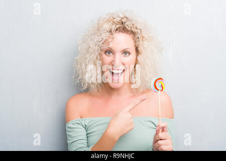 Young blonde woman over grunge grey wall eating candy lollipop very happy pointing with hand and finger Stock Photo