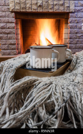 Big enameled mugs with tea, and warm wool scarf , opened book , near cozy fireplace, in country house, winter vacation, vertical. Stock Photo