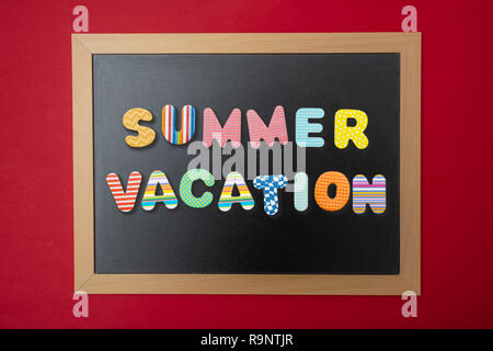 Summer vacation concept. Black chalkboard with wooden frame, text summer vacation in colorful letters, red wall background Stock Photo