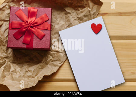 Red box with a gift and next to a white blank sheet for text with a heart on a wooden surface. Festive concept for Valentine's Day or Women's Day or Mother's Day. Stock Photo