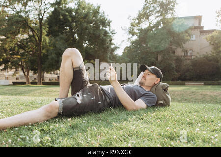 A young guy with a backpack or a student is lying on the grass during a break or is resting and using a tablet. He is watching a video or reading or something else. Stock Photo