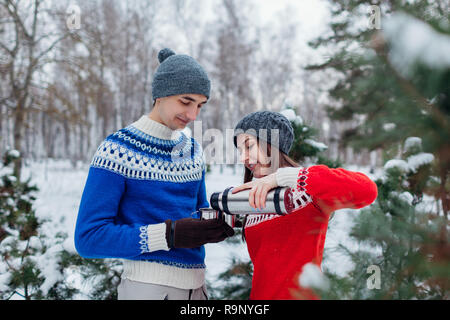 Young couple pouring hot tea out of thermos into cups in winter forest. Happy people relaxing outdoors during holidays Stock Photo