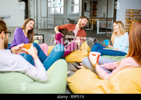 Group of a young coworkers having fun sitting on the colorful poufs playing guitar during the coffee break in the office Stock Photo