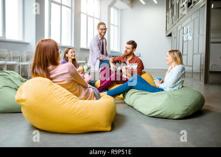 Group of a young coworkers having fun sitting on the colorful poufs playing guitar during the coffee break in the office Stock Photo