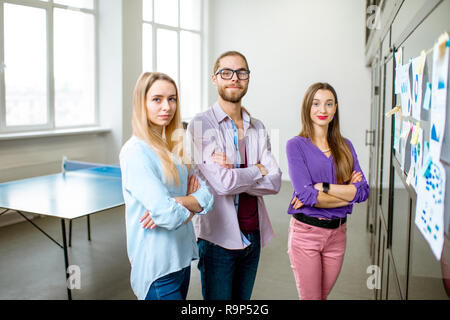 Portrait of a young team of coworkers dressed casually standing near the glass wall with some statistics and stickers in the office Stock Photo