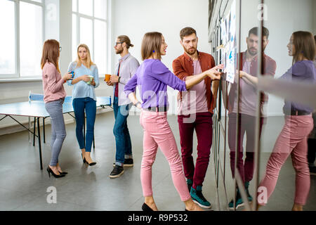 Young team of coworkers dressed casually talking near the glass wall with some statistics and stickers in the office Stock Photo