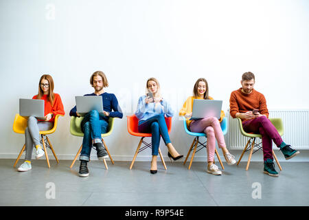 Young coworkers working with gadgets sitting in a row on the colorful chairs on the white wall background indoors Stock Photo