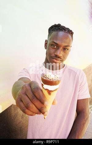 Young handsome black man wears a pink t-shirt, holds and eats a ice cream cone in summertime on a painted wall as a sunrise or sunny day Stock Photo