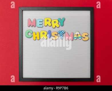 Merry Christmas. Board with black frame, text Merry Christmas in colorful letters, red wall background, copy space Stock Photo