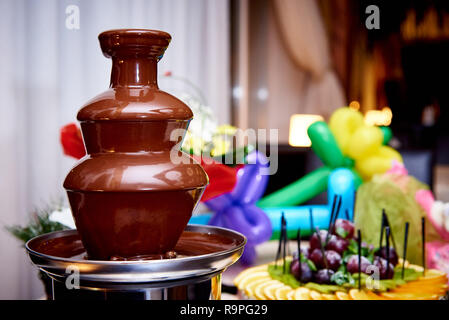 Chocolate fountain on a blurred background with fresh fruits and gifts. Stock Photo