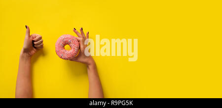 Female hand holding pink donut and showing like, ok sign over yellow background. Top view, flat lay. Diet, calories, healthcare, willpower, summer, bo Stock Photo