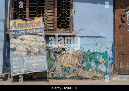 Scruffy, faded and wonky Beauty Parlour A-Board propped against a traditionally painted wall featuring a fight scene with elephants.  Bundi, Rajasthan Stock Photo