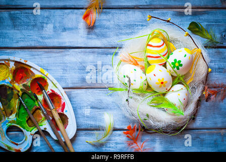 Handcrafted Easter eggs in the nest and palette with bbrushes and paints on wooden background. Stock Photo