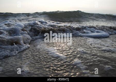 Small waves lap a beach as the tide goes out, curling up the sand and making dirty bubbles mixed with clean white foam, as larger waves build in the b Stock Photo