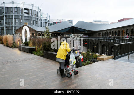 Rear view of a woman and pushchair with shopping bags and a view Gasholders apartment building and Coal Drops Yard Kings Cross London UK  KATHY DEWITT