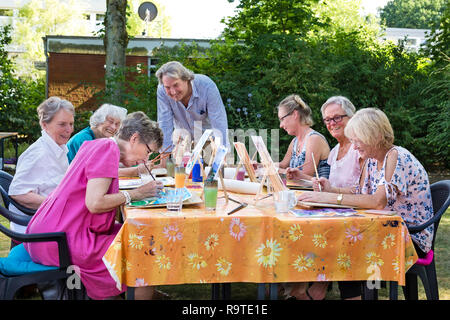 Male artist giving art lessons to the group of senior women, practicing in painting pictures sitting at one table outdoors in backyard. Stock Photo