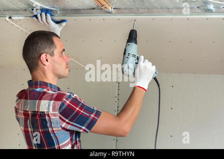 Young man in usual clothing and work gloves fixing drywall suspended ceiling to metal frame using electrical screwdriver on ceiling insulated with shi Stock Photo