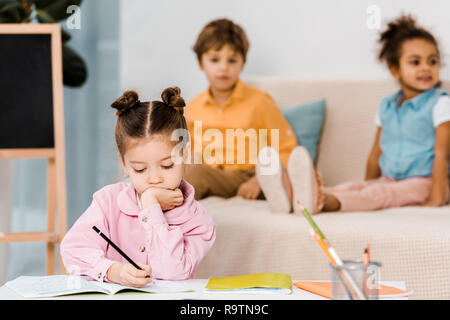 adorable little child writing with pencil while friends sitting behind Stock Photo