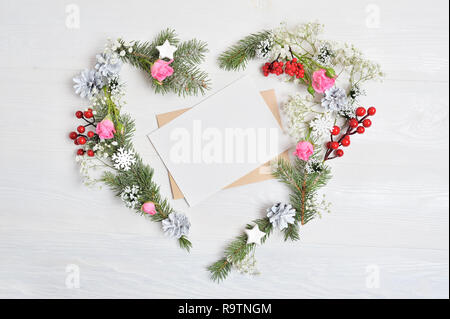 Mockup of Christmas Wreath in form of heart with sheet of paper Decorated with white snowflakes and cones. White wooden background with place for your text Stock Photo
