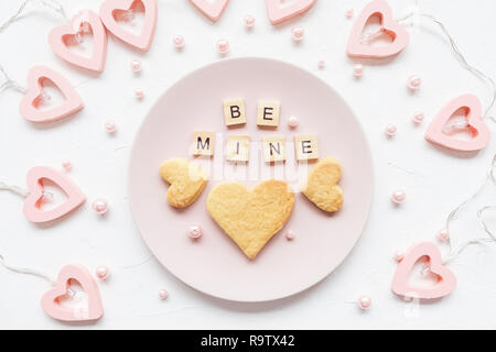 BE MINE words and heart shaped butter cookies on a pink plate and Valentine's Day decoration - heart shaped lights for a Valentines Day on a white bac Stock Photo