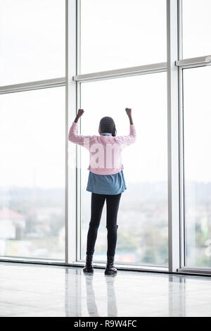 Full height afro american woman with reised hands up standing in front of pamoramic window of celebrate success. Rear view. Stock Photo