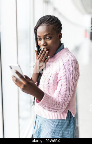 Young surprised afro woman read shocked news in internet from phone in front of panoramic windows Stock Photo