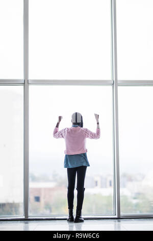 Full height afro american woman with reised hands up standing in front of pamoramic window of business center celebrate success. Rear view. Stock Photo