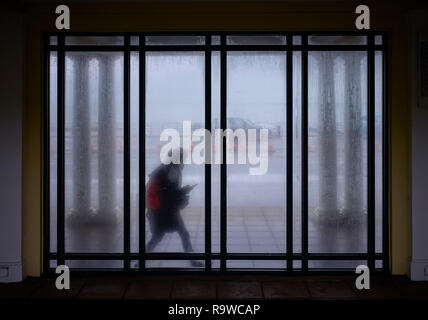 Young woman looking at mobile phone while walking past raindrop patterened windows in Douglas, Isle of Man Stock Photo