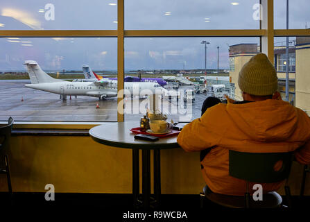 Man wearing woollen hat looking out from Douglas airport cafe at the planes on the tarmac below