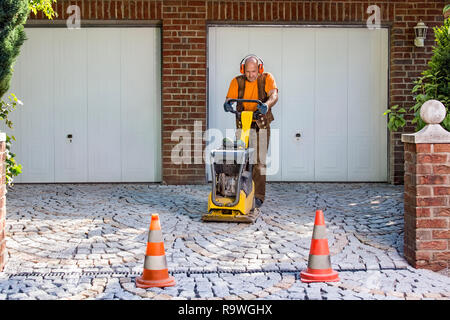Builder or contractor laying new paving bricks in a house driveway using a mechanical compacter for compaction of the cement pavers. Stock Photo