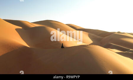 Aerial view from a drone flying next to a woman in abaya (United Arab Emirates traditional dress) walking on the dunes in the desert of the Empty Quar Stock Photo