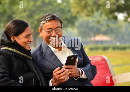Senior couple working on mobile and talking, smiling, laughing  in a park in winters in Delhi, India Stock Photo