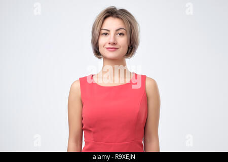Charismatic attractive woman in red dress, smiling cheerfully while standing against gray background Stock Photo