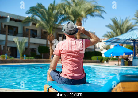 Young Man in Hat Sitting On Sun Loungers By Swimming Pool hotel Stock Photo