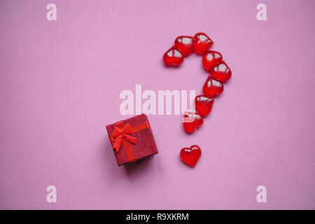 A box with a gift and a number of question marks laid out from small red hearts. Concept for Valentine's Day or another love event. A question mark means a surprise. Stock Photo