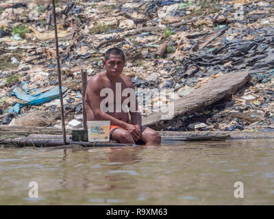 Iquitos, Peru - Sep 25, 2018: The man sits on the banks of the Itaya River and cools his body with water. A huge pollution can be seen in the backgrou Stock Photo