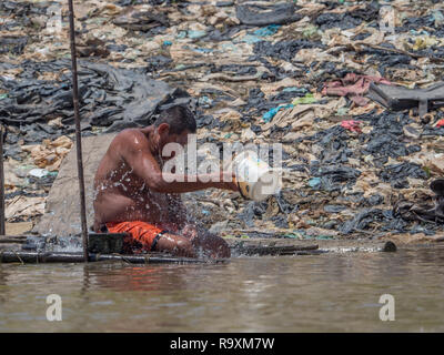 Iquitos, Peru - Sep 25, 2018: The man sits on the banks of the Itaya River and cools his body with water. A huge pollution can be seen in the backgrou Stock Photo