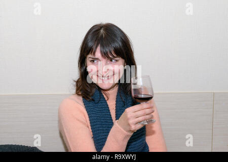 Close-up head and shoulder portrait of smiling brunette mature adult woman with glass of wine in her hand on light wallpaper background. Stock Photo