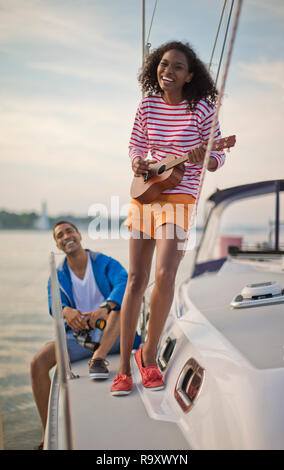 Portrait of a smiling young woman having fun playing the ukulele while sailing. Stock Photo