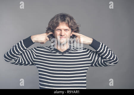 Displeased man covering ears with fingers doesn't want to listen anything isolated on gray background. Young handsome male squinted his eyes looking a Stock Photo