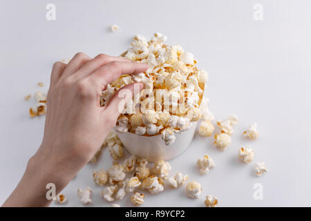 A hand is taking popcorn from a full paper bucket on a white background. Top view. Close-up. Stock Photo