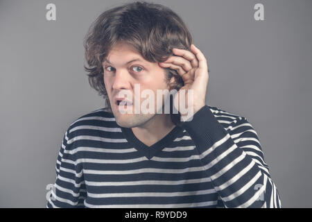 Closeup of curious or deaf man placing hand on ear asking someone to speak up or listening to bad news, isolated on grey background. Negative emotion  Stock Photo